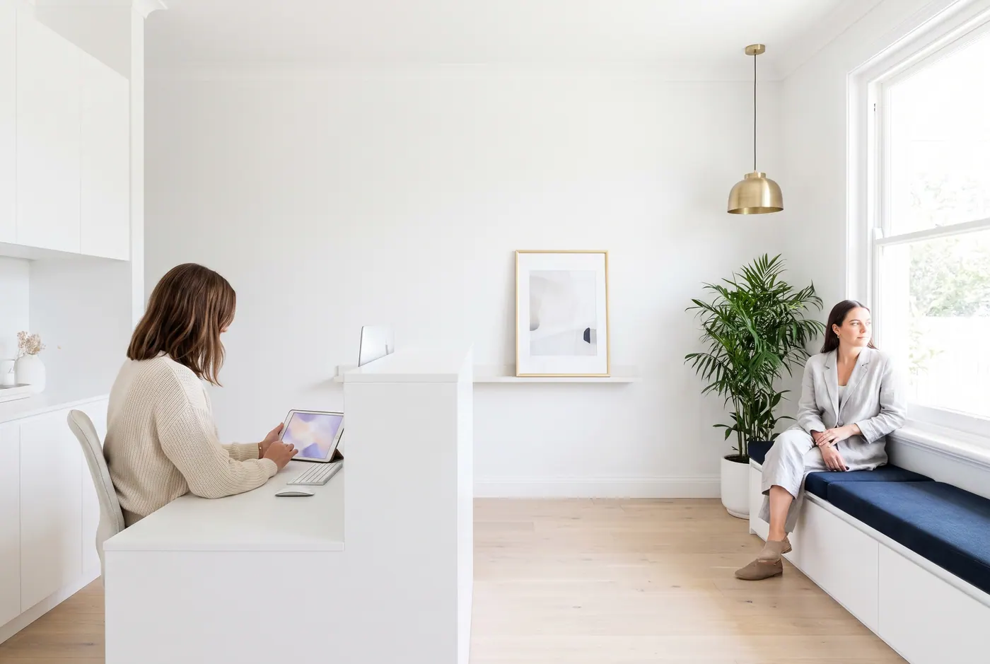 A receptionist at a bright, white clinic front desk checking a tablet while a patient waits on a window seat.